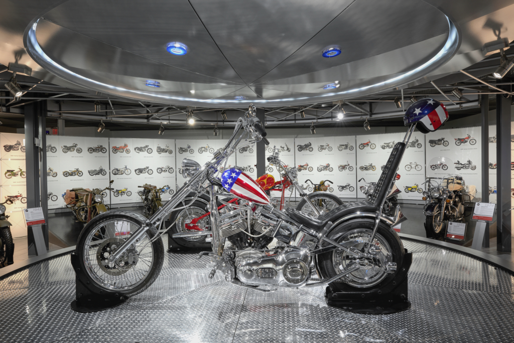 Showcase of a custom chopper motorcycle with a patriotic American flag design on the fuel tank and helmet at the Deeley Motorcycle Exhibition. The display is set on a polished metal platform under modern lighting, surrounded by a wall of historical motorcycle images and additional vintage bikes in the background.