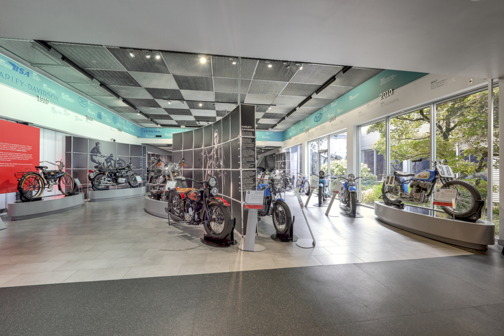 Wide-angle view of the Deeley Motorcycle Exhibition hall showcasing an extensive collection of vintage and classic motorcycles. The display includes various models from different eras, informational panels, and a timeline above the exhibits marking significant events in motorcycle history from 1910 to 2010. Large windows provide natural light and a view of the garden outside.