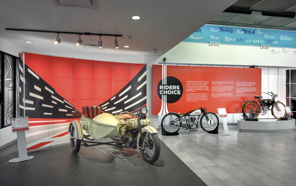 Exhibit at the Deeley Motorcycle Exhibition featuring a variety of historical motorcycles, including a 1928 model with a sidecar. The display includes a 'Riders Choice' section with informational panels and a visually striking red and black backdrop. The timeline on the wall highlights key events in motorcycle history from 1900 to 1910.